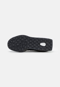 Suela de zapato deportivo negro con goma texturizada, que presenta patrones circulares y ovalados para mayor agarre y una zona del talón lisa. Diseño de punta redondeada.