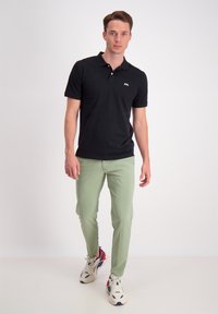 Polo nero con maniche corte e colletto, abbinato a pantaloni verde chiaro. Include un dettaglio con logo sulla maglietta. Sfondo bianco.