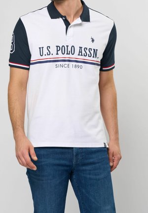 Mężczyzna ubrany w biały polo z granatowymi rękawami, logo i napisem U.S. Polo Assn., w połączeniu z niebieskimi dżinsami, stoi na jednolitym tle.