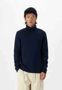 Suéter de cuello alto de punto en color navy con un patrón texturizado, mangas largas y puños de canalé, combinado con pantalones de color beige claro.