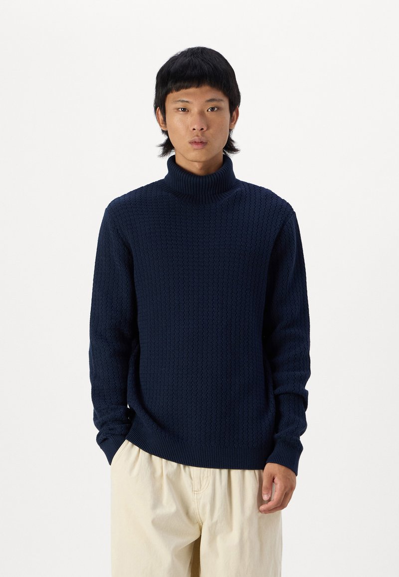 Suéter de cuello alto de punto en color navy con un patrón texturizado, mangas largas y puños de canalé, combinado con pantalones de color beige claro.