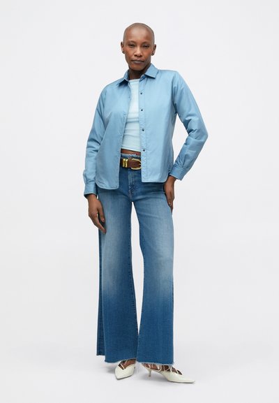 Lichtblauw button-up shirt, aansluitende mintkleurige tanktop, high-waisted flared jeans en crèmekleurige puntige hakken. Gestructureerde denim en glanzende riem.