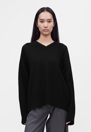 Jeune femme aux longs cheveux noirs portant un pull ample noir à col en V et un pantalon gris, debout devant un fond blanc.