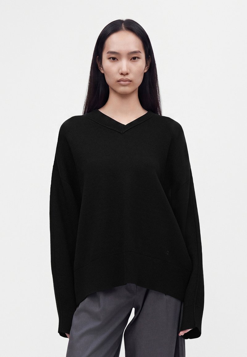 Jeune femme aux longs cheveux noirs portant un pull ample noir à col en V et un pantalon gris, debout devant un fond blanc.