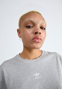 Pelēka kokvilnas kreklu krekls ar izšūtu baltu Adidas logo uz krūtīm. Tuvplāns ar personu ar īsiem platīna blondiem matiem un riņķveida auskariem.
