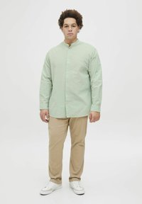 Camisa de manga larga verde claro con cuello de banda y cierre de botones, combinada con pantalones beiges y zapatillas blancas, de pie sobre un fondo neutro.
