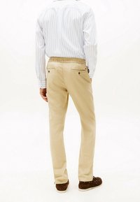 Beige Hosen mit lockerer Passform, elastischem Bund und zwei Gesäßtaschen. Kombiniert mit einem hellblauen, senkrecht gestreiften Hemd.