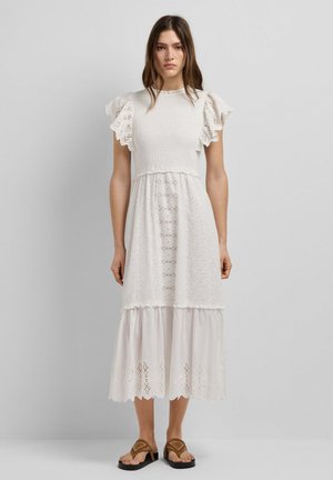 Vrouw draagt een witte midi-jurk met gerimpelde korte mouwen, oogjespatronen en bruine sandalen met platte zolen, staande tegen een effen grijze achtergrond.