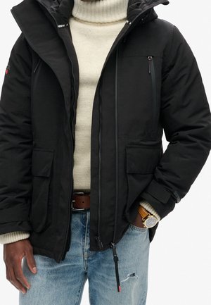 Mann trägt schwarze Winterjacke mit Kapuze über beigem Rollkragenpullover, blaue zerrissene Jeans und eine braune Lederarmbanduhr am linken Handgelenk.