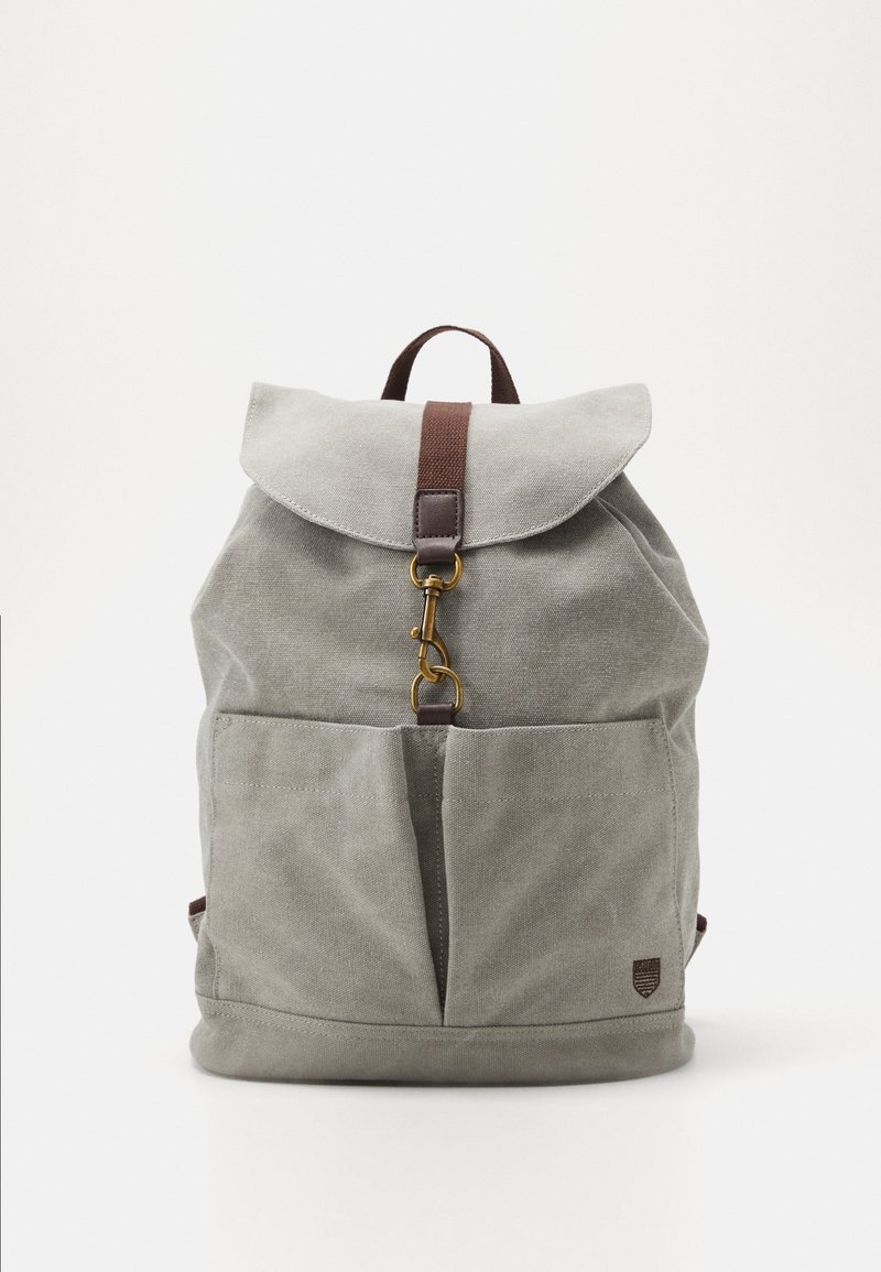 Sac à dos en toile grise avec poches avant, sangle marron et fermeture à crochet en laiton sur fond blanc uni.