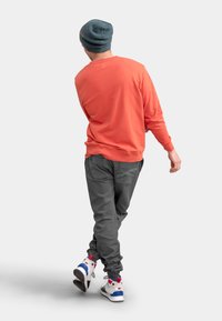 Oranje sweatshirt, grijze broek met zichtbare zakken, en witte sneakers met rode en blauwe accenten. Casual, relaxte pasvorm en textuur.