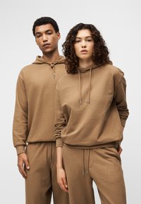 Δύο μοντέλα φορούν ταιριαστά καφέ φούτερ με κουκούλα και παντελόνια. Το ύφασμα φαίνεται μαλακό με υφή. Τα κορδόνια έχουν ασημένια άκρα.