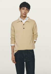 Pullover beige con colletto e tre bottoni neri, indossato sopra una camicia bianca. Jeans in denim scuro, tessuto a maglia testurizzata, vestibilità rilassata.