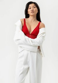 Bralette rosso in pizzo con bordi scallop e scollatura profonda, indossato sotto un blazer bianco oversize e pantaloni abbinati. Texture di tessuto liscia.