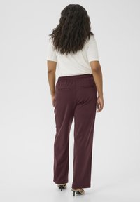 Burgundy trousers with a relaxed fit, featuring an elastic waistband, two back pockets, and smooth fabric. Worn with a white top.