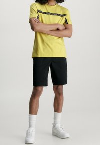 Žluté bavlněné tričko s černým potiskem loga na hrudi, v kombinaci s černými šortkami a bílými teniskami.