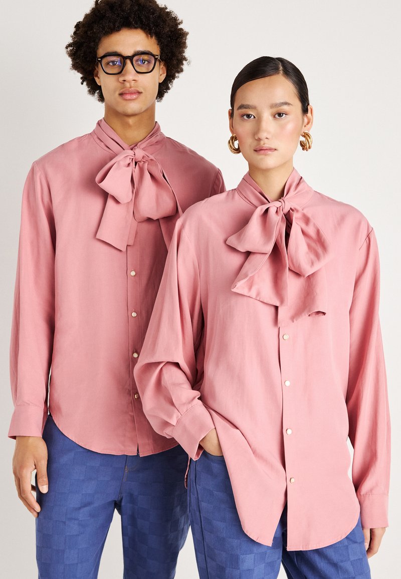 Dois modelos usam camisas de manga longa cor-de-rosa claro com grandes gravatas-borboleta no pescoço. As camisas são de tecido suave e têm fechos de botão. Um dos modelos combina com calças azuis estampadas.