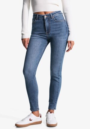Frau trägt blaue High-Waist Skinny Jeans, weißen kurzen gerippten Pullover und weiße Sneaker mit braunen Akzenten, steht vor weißem Hintergrund.