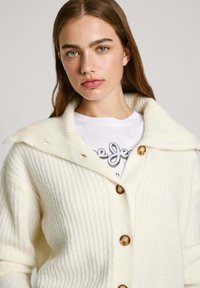 Romige geribbelde cardigan met grote kraag, voorzien van bruine knopen, gedragen over een wit graphic t-shirt. Zachte textuur, casual ontwerp.