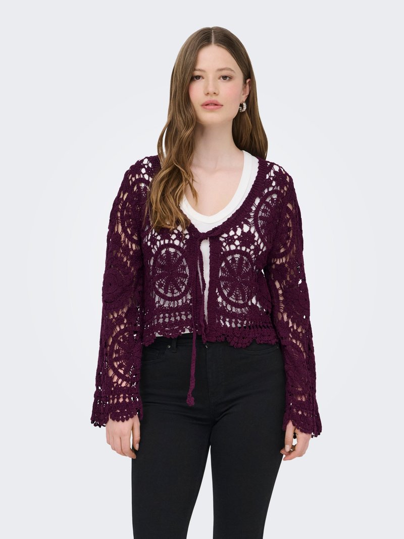 Jeune femme portant un cardigan en crochet violet par-dessus un haut blanc et un pantalon noir, debout devant un fond clair uni.