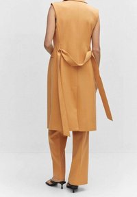 Bez rękawów, pomarańczowa kamizelka o dopasowanym kroju z tylnym sznurkiem, w połączeniu z dopasowanymi spodniami slim fit i czarnymi szpilkami, na neutralnym tle.