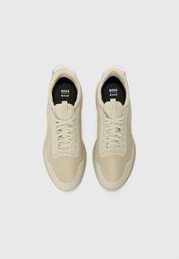 Beige athletic shoes with a textured upper, lace closure, and rounded toe. Features branding on the tongue and cushioned sole.