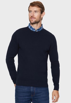 Pull en maille marine à texture côtelée, col rond et manches longues, porté par-dessus une chemise à col à carreaux bleue, associé à un jean.
