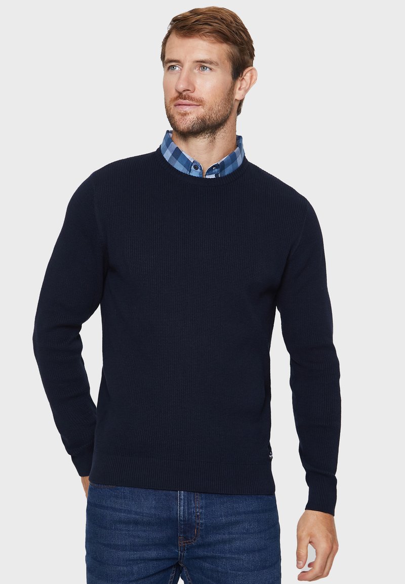 Marineblauer Strickpullover mit gerippter Textur, Rundhalsausschnitt und langen Ärmeln, getragen über einem blau karierten Hemd mit Kragen, kombiniert mit Jeans.