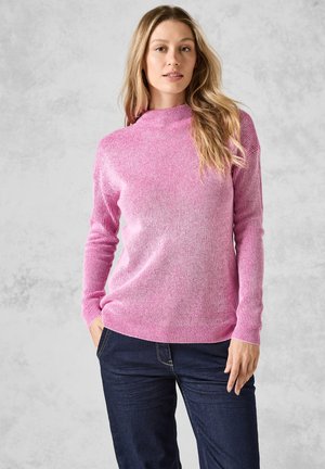 Roze ribgebreide trui met lange mouwen en een hoge halslijn, met variaties in textuur. Combinatie met donkerblauwe jeans. Geen zichtbare logo's of patronen.