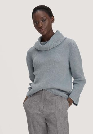 Vrouw draagt een lichtblauwe gebreide trui met col en een grijze broek met visgraatmotief, handen in de zakken.