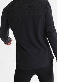 Hombre vistiendo una camiseta atlética de manga larga negra con un panel posterior texturizado y leggins atléticos negros a juego, posando frente a un fondo blanco.
