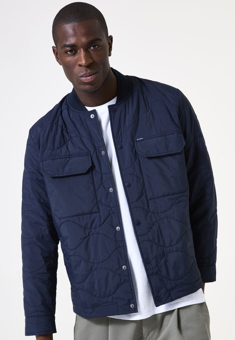 Marineblauer gesteppter Blazer mit Druckknopfverschluss, zwei Fronttaschen und einem gerippten Kragen, über einem weißen Hemd getragen.