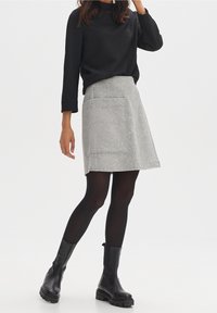 Grauer, texturierter A-Linien-Rock mit großer Vorderstasche, kombiniert mit einem schwarzen Langarmshirt und schwarzen Stiefeletten.