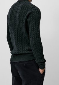 Dunkelgrüner gestrickter Pullover mit einem Kabelmuster, gerippten Bündchen und Saum, kombiniert mit dunklen Hosen. Blick von hinten.