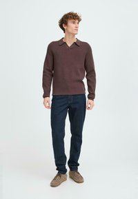 Pulover tricotat maro cu decolteu în V și guler, combinat cu blugi de denim închisi la culoare și pantofi din nubuc maro deschis, stând pe un fundal deschis.