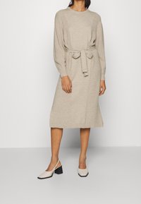 Robe beige tricotée longueur genoux avec ceinture à nouer à la taille, munie de manches longues et d'une texture côtelée, associée à des chaussures à talons de couleur claire.