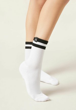 Person's lower legs wearing white ribbed crew socks with black double stripes and a small black tag near the cuff on a light background.