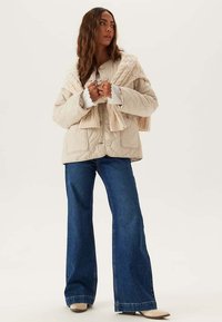 Beige gewatteerde jas met grote knopen, geribde manchetten en gestructureerde accenten, gecombineerd met licht uitlopende blauwe jeans en lichtbeige schoenen.