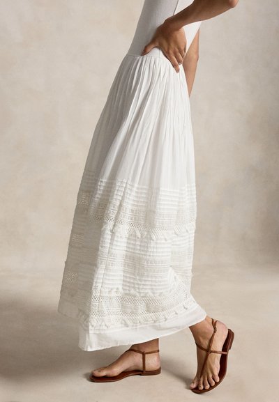 Naine, kes kannab valget plisseeritud maksiseelikut pitsdetailidega ja pruune madalaid sandaale, seisab ühe käega puusal.