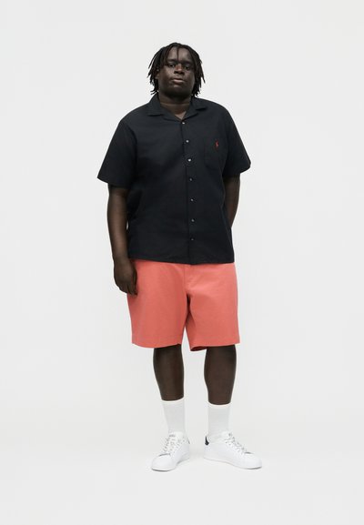 Mężczyzna z dredami stojący, ubrany w czarną koszulę na krótki rękaw zapinaną na guziki, różowe szorty, białe skarpetki i białe trampki na jednolitym tle.