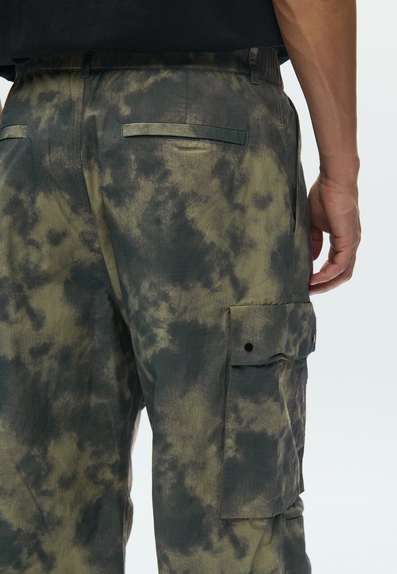 Pantalon cargo en vert olive avec un motif tie-dye gris foncé, doté de deux poches arrière et d'une poche cargo latérale avec fermeture à pression.