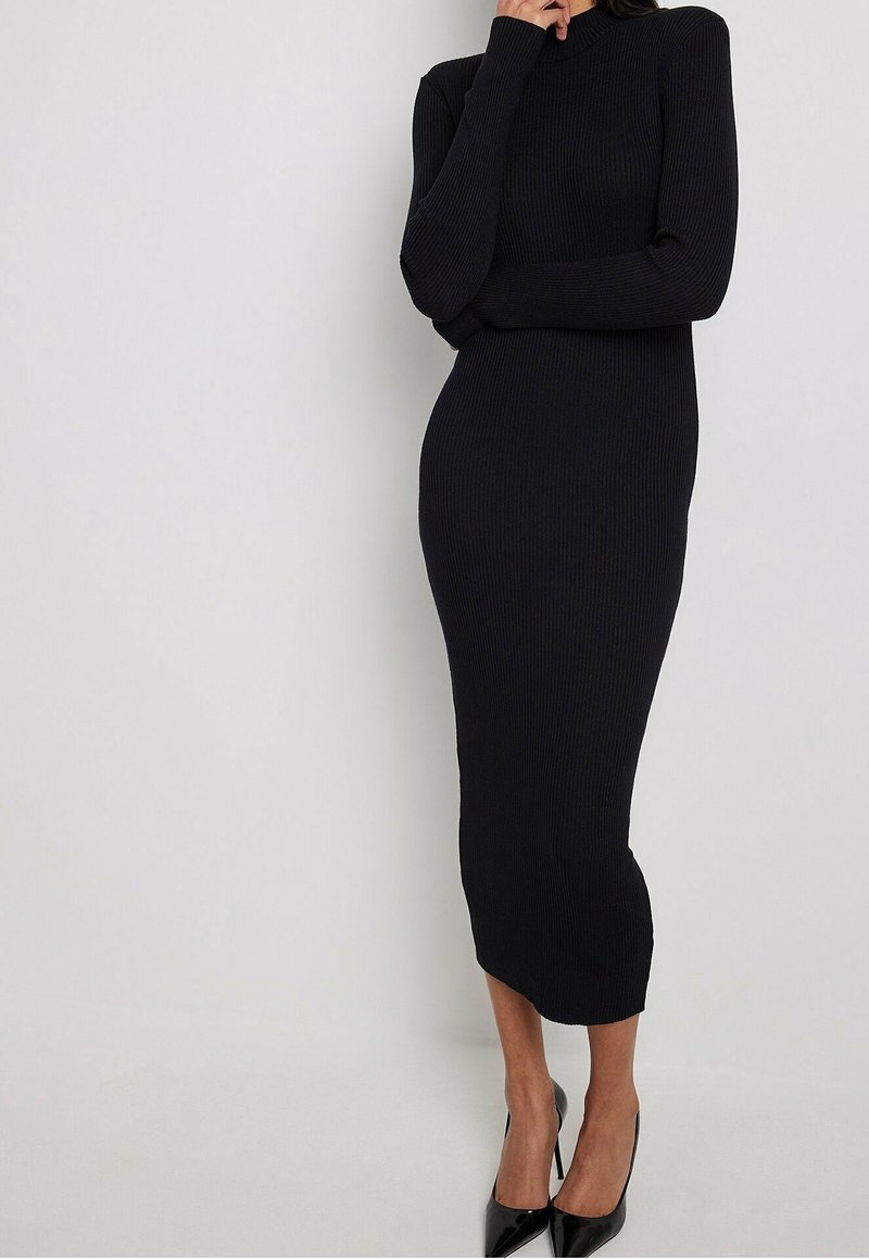 Robe en maille noire côtelée avec manches longues, col haut et coupe ajustée, atteignant le milieu du mollet. Présente une texture lisse et un design minimaliste.