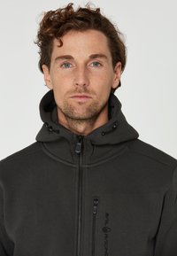 Dunkelgraue Kapuzenjacke mit durchgehendem Reißverschluss, strukturiertem Stoff und einer kleinen Brusttasche, die links mit einem schwarzen Logo versehen ist.