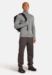 Uomo che indossa una giacca grigia con zip, pantaloni scuri, scarpe da trekking, un berretto arancione e porta uno zaino nero a spalla.