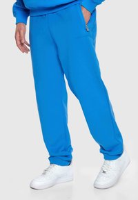 Blaue Sweatpants haben eine glatte Textur, einen elastischen Bund, eine seitliche Reißverschlusstasche und werden mit weißen Sneakers kombiniert.