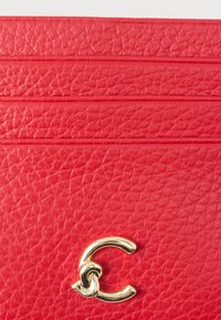 Nærbillede af rød tekstureret læder med vandret syninger og et lille skinnende guldlås i form af et sammenknyttet "C".