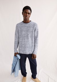 Helles graues strukturiertes Strickpullover mit langen Ärmeln, marineblaue Hose und beigefarbene Slipper. Hält eine Jeansjacke in der linken Hand.