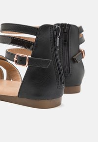 Mustat keinonahkasaappaat, joissa on useita hihnoja, takaosassa vetoketjukiinnitys ja ruskea teksturoitu pohja.