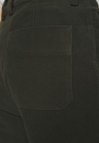 Dark green pants featuring a back pocket with a rectangular shape. The fabric appears smooth with subtle texture and contrast stitching.