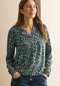 Langsleeve blouse in donker groen met een abstract dierenprint ontwerp, voorzien van een kraag en knopenlijst, gecombineerd met blauwe denim jeans.